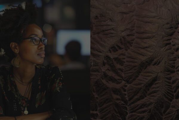 A woman with glasses and large earrings sits attentively in a dimly lit environment, with a blurred background of people and