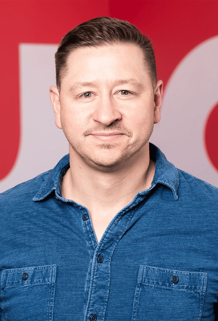 A man with short dark hair, light skin, and a slight beard, wearing a blue denim shirt, smiles in front of a red background.