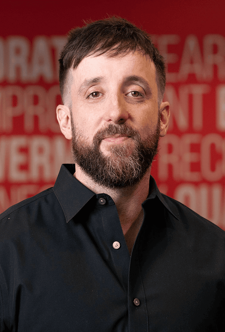 A man with short brown hair, a beard, and mustache wearing a black collared shirt stands in front of a red background with