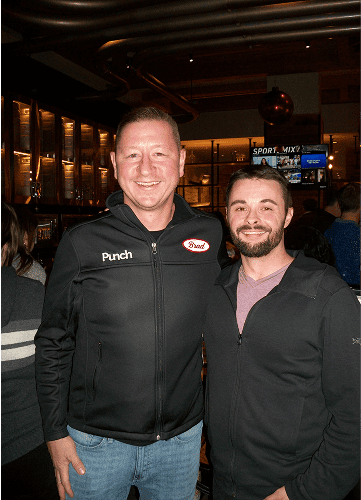 Two smiling men stand together in a bar or restaurant, one wearing a black jacket with "Punch" and "Dale" logos.