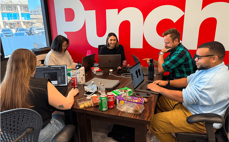 Five people are gathered around a table with snacks and drinks, working on laptops in a room with a red wall and large