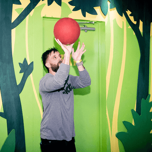 Punch Team Culture Photo 15 A man with a beard and gray sweatshirt reaches up to catch a large red ball against a vibrant green and jungle-themed