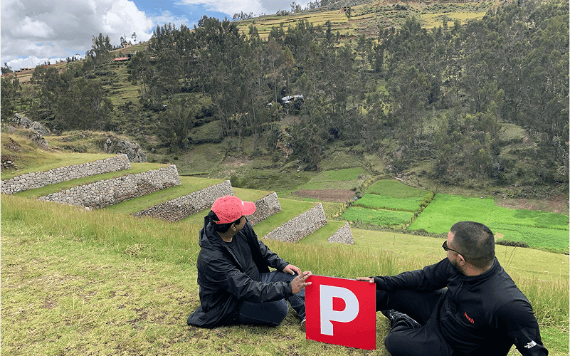 Two people sit on a grassy hillside holding a red sign with a white "P" against a backdrop of terraced fields and a forested