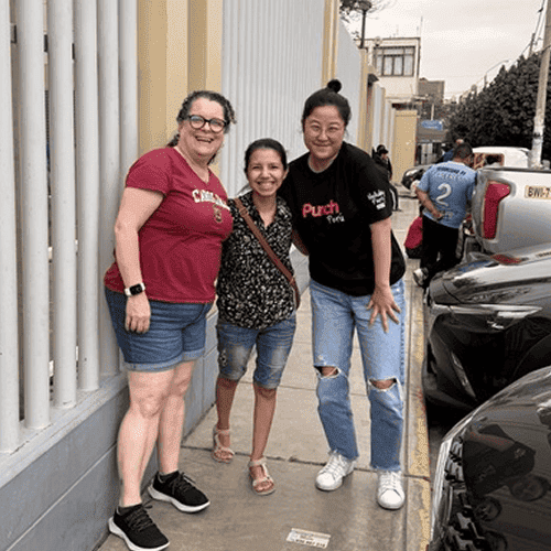 Punch Team Culture Photo 18 Three women stand together smiling on a sidewalk, with parked cars and a building in the background.