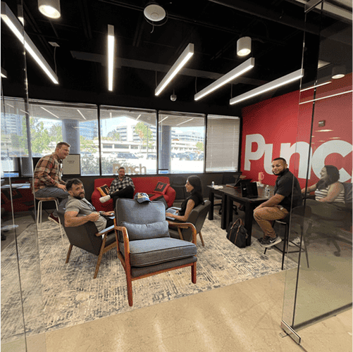 Six people are seated and standing in a modern office lounge with large windows, a red wall with "Punc" written on it, and