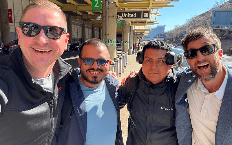 Four men stand together outdoors at an airport, smiling and posing for a selfie with luggage and airport signs in the