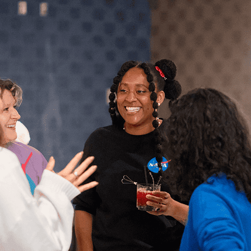 Punch Team Culture Photo 7 A woman with braided hair and a NASA shirt smiles while holding a drink, engaging with two other women in conversation.